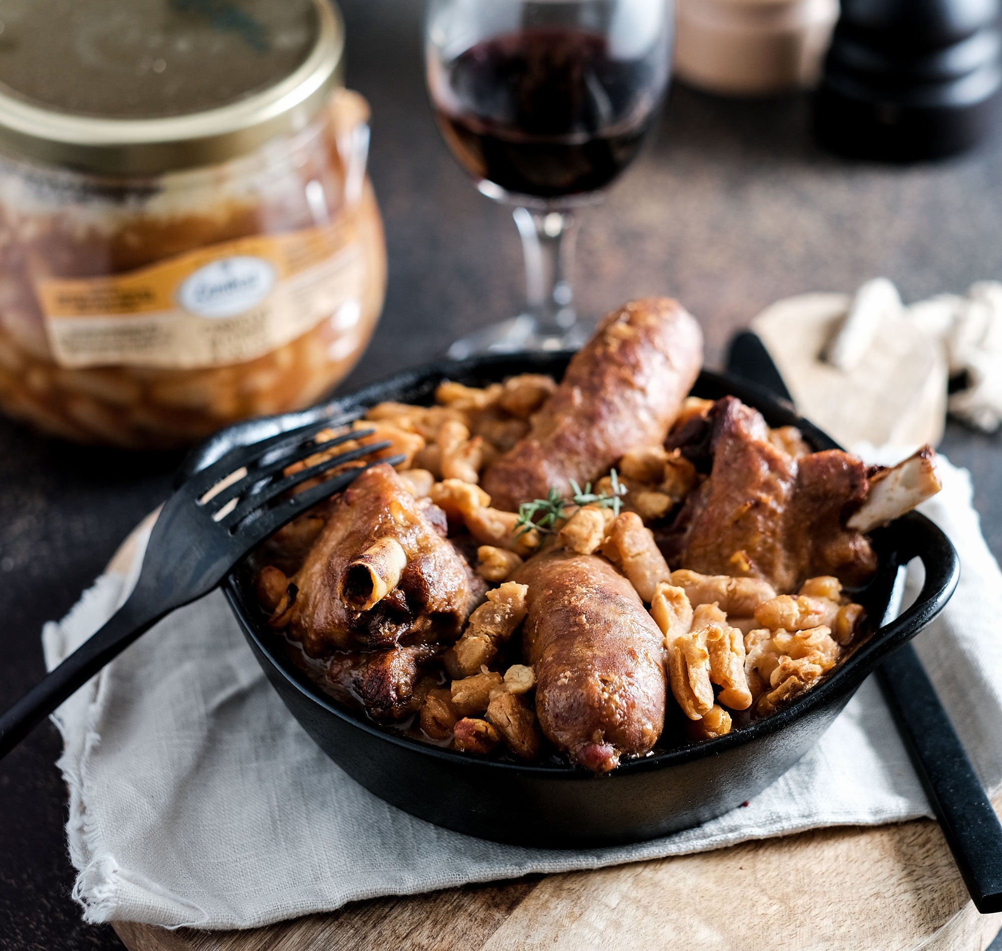 Cassoulet de canard au confit servi dans un plat noir avec notre cassoulet en bocal et un verre de vin rouge