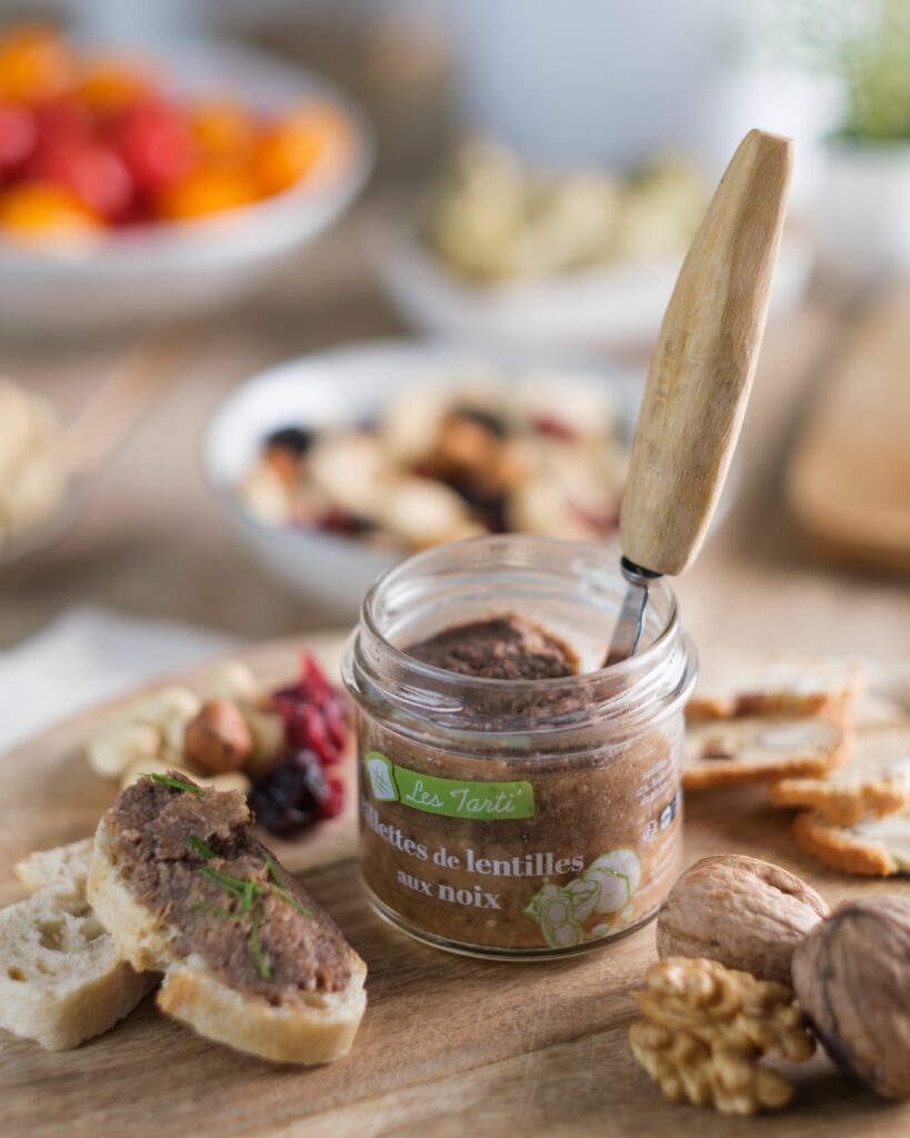 Tartinable apéro : tarti rillettes de lentilles aux noix du Périgord ouvert avec couteau dans le bocal, tartine garnie et tranches de pain sur une planche en bois