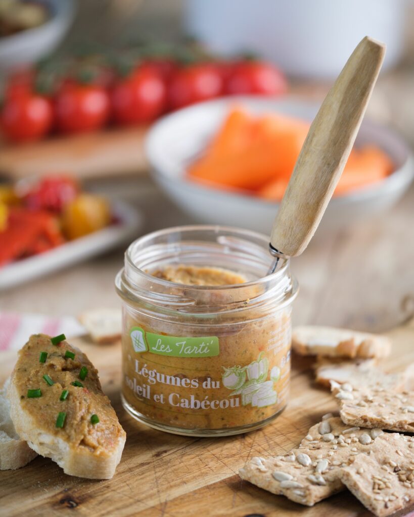Tartinables apéro : bocal de tarti légumes soleil et cabécou ouvert, présenté sur une planche en bois avec une tartine gourmande à côté.