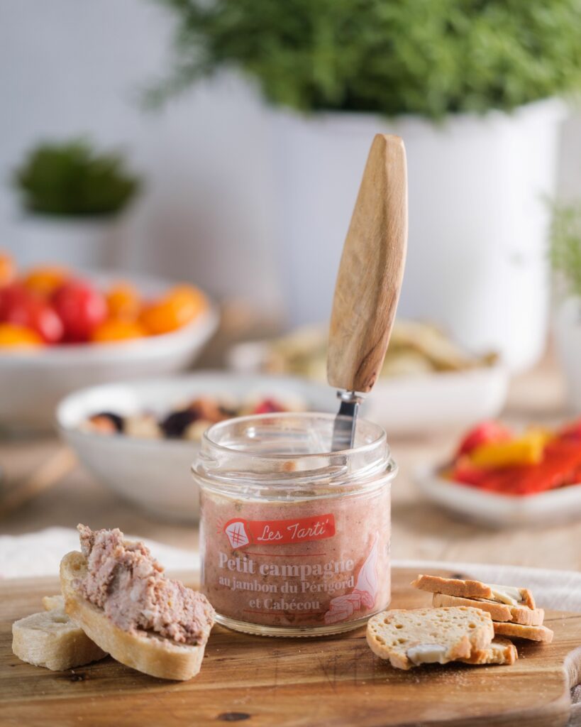 Bocal de tarti petit campagne jambon du Périgord et cabécou ouvert, présenté sur une planche en bois avec une tartine gourmande à côté.