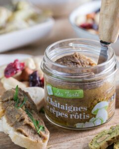 Bocal de tarti châtaignes et cèpes rôtis ouvert, présenté sur une planche en bois avec une tartine gourmande à côté.