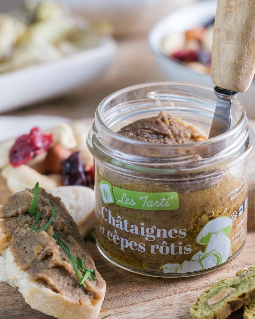 Tartinable apéro : bocal de tarti châtaignes et cèpes rôtis ouvert, présenté sur une planche en bois avec une tartine gourmande à côté.