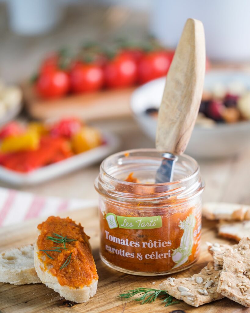 Tartinables apéro : tarti tomates rôties carottes céleri Maison Lembert dans un bocal ouvert avec une tartine garnie et des légumes frais en arrière-plan