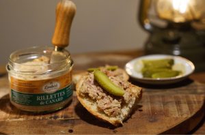 Rillettes de canard artisanales Maison Lembert sur tranche de pain de campagne, accompagnées d’un cornichon, avec le pot de rillettes en arrière-plan.