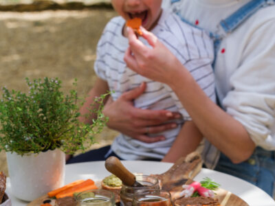 Idées apéritif été : enfant dégustant un tartinable sur les genoux de sa maman lors d’un apéritif d’été convivial avec des tartinables artisanaux colorés