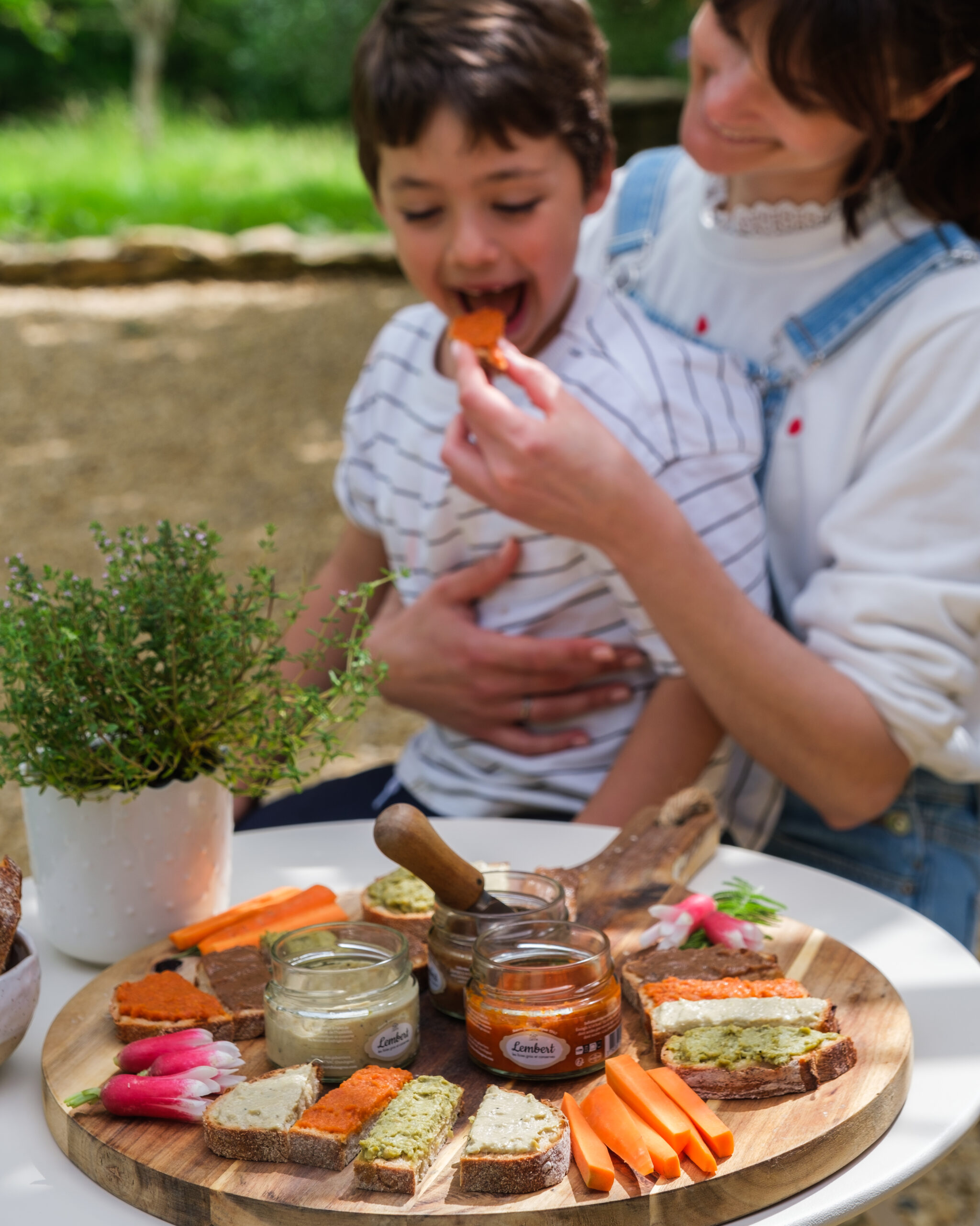 Idées apéritif été : enfant dégustant un tartinable sur les genoux de sa maman lors d’un apéritif d’été convivial avec des tartinables artisanaux colorés