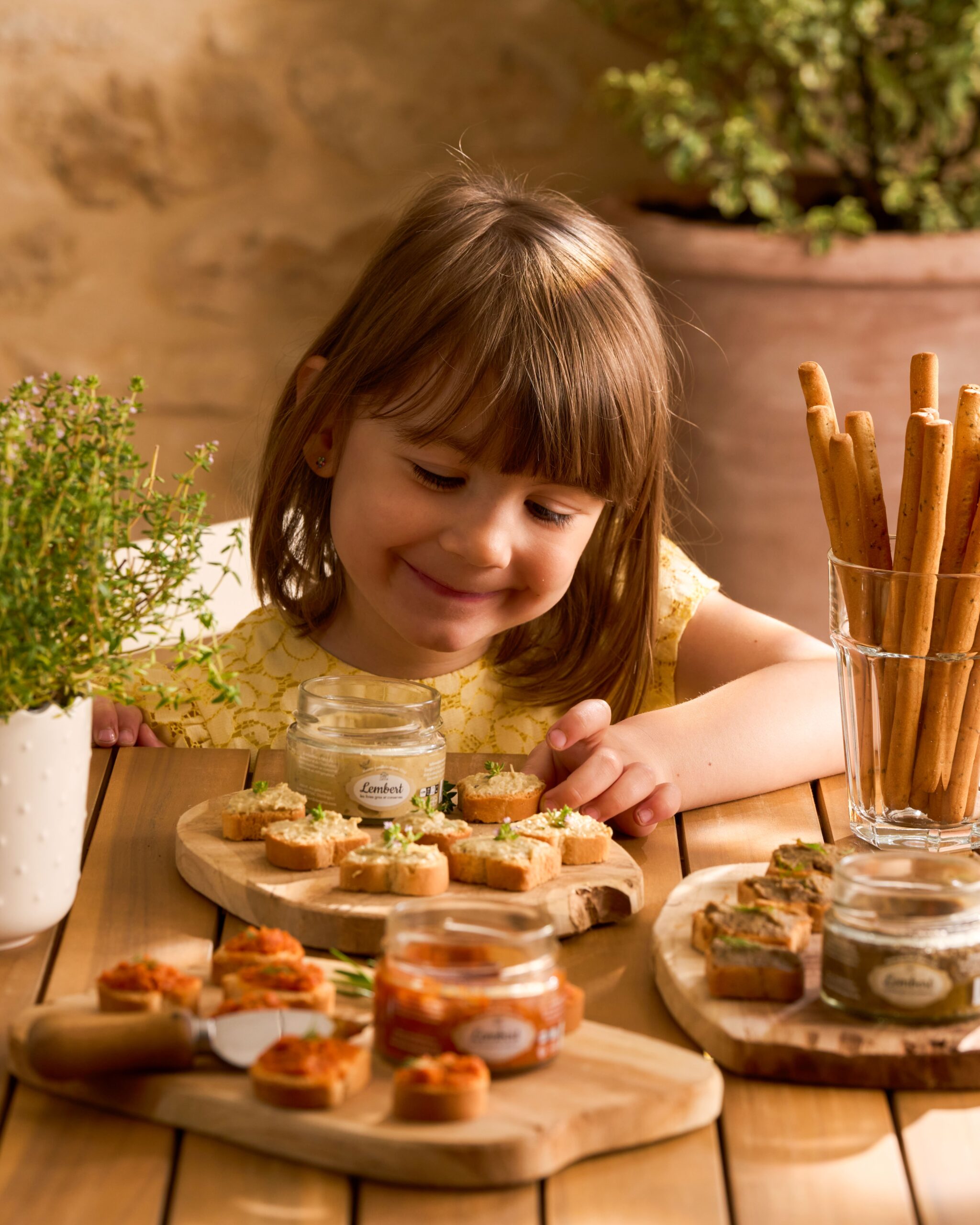 Petite fille souriante devant des tartinables apéro artisanaux sur planches en bois avec gressins